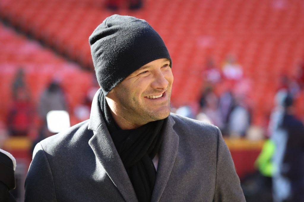 CBS broadcaster Tony Romo smiles while wearing a black beanie, grey coat, and black scarf.