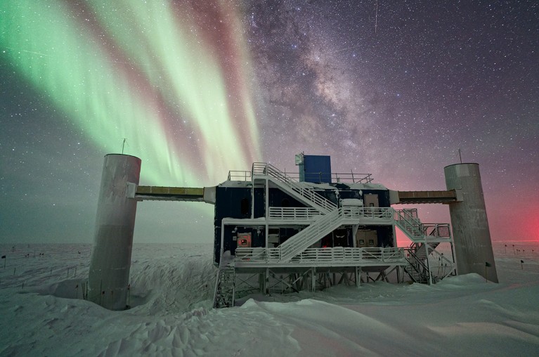 The IceCube Lab is seen under a starry, night sky, with the Milky Way appearing next to striking auroras in the background.