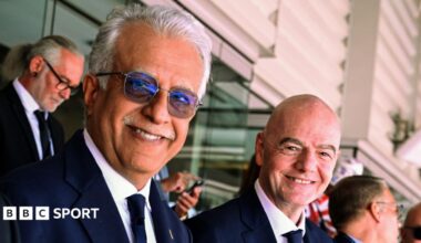 Sheikh Salman bin Ibrahim Al Khalifa and Gianni Infantino stood next to one another in the stands of a football stadium, smiling and looking to their right towards the camera