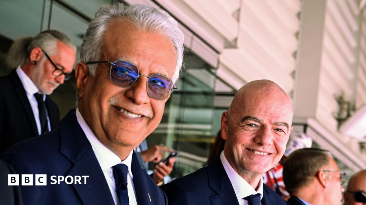 Sheikh Salman bin Ibrahim Al Khalifa and Gianni Infantino stood next to one another in the stands of a football stadium, smiling and looking to their right towards the camera
