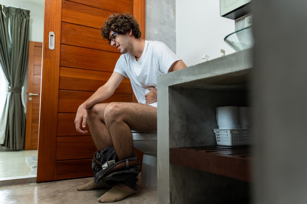 A young man sitting on a toilet in a bathroom, holding his stomach in pain.