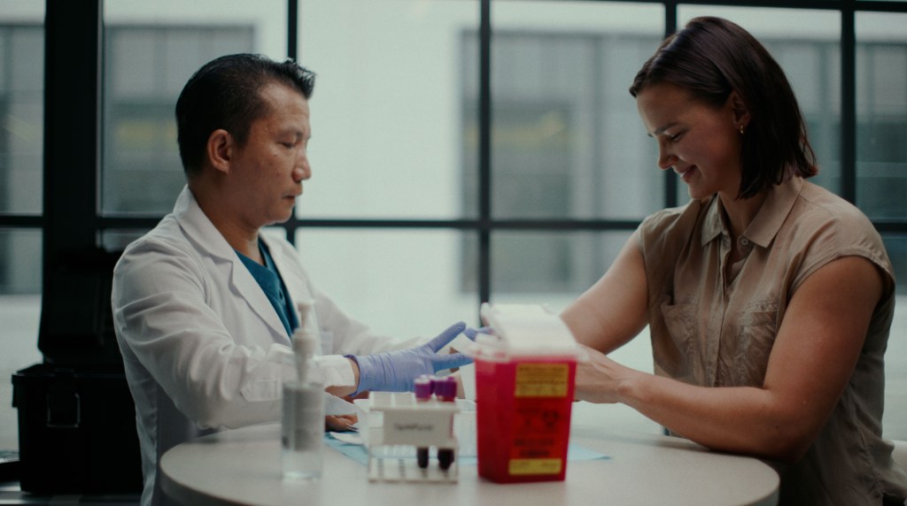A male doctor in a white coat and blue gloves prepares to draw blood from a smiling female patient seated across from him at a table.