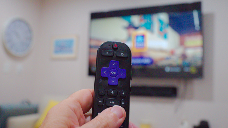 Close-up of a hand holding a Roku remote with a TV in the background.