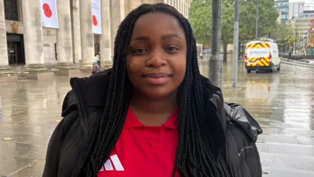 Chenai Masunungure wearing a red Manchester United top and a black coat. She's stood in Manchester city centre smiling for the camera with long black braided hair.