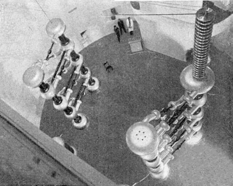 A black and white photo of two ladder-like stacks of components that control charge, topped by capacitators, which store charge. Between them are the vacuum tubes the charges move through