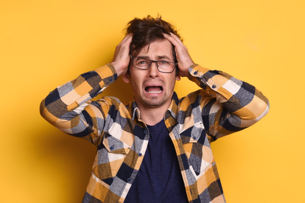 A stressed young man in glasses and a plaid shirt holds his head in despair against a yellow background.