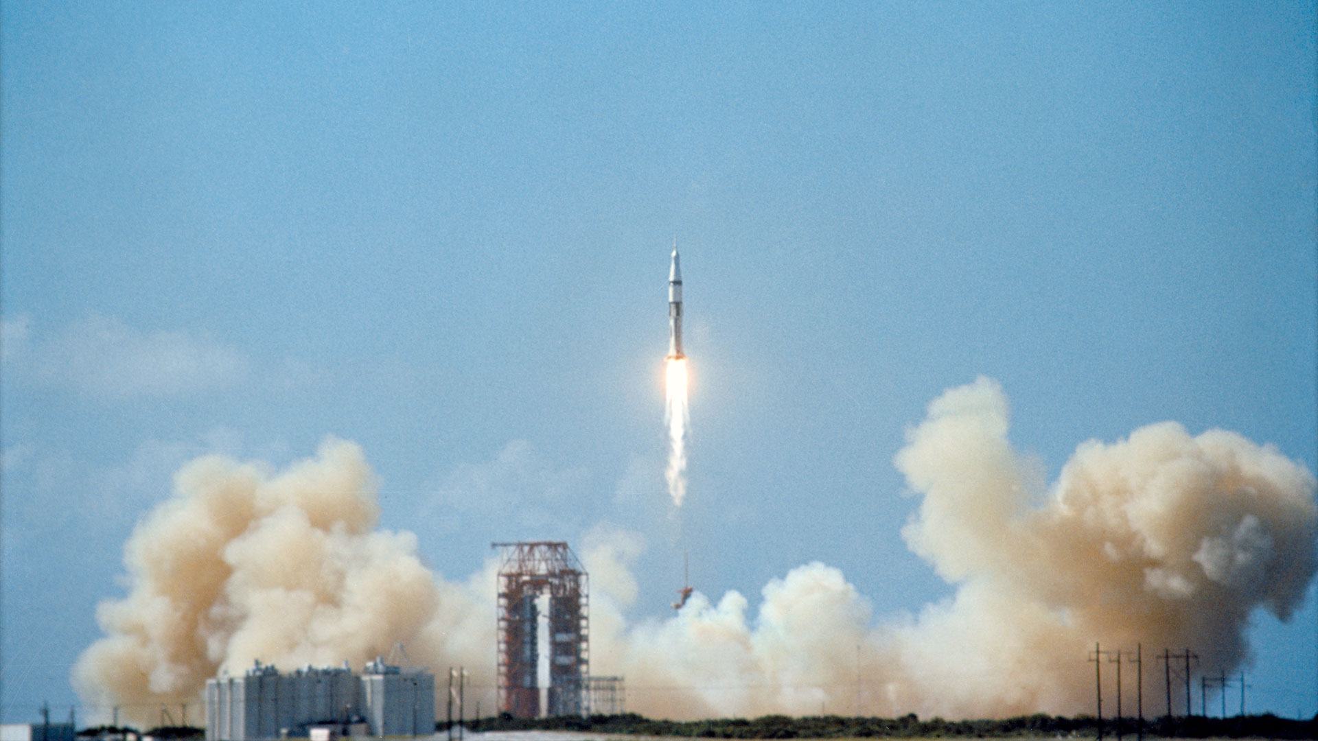 a white and black rocket lifts off into a blue sky leaving behind a plume spread across its launch site