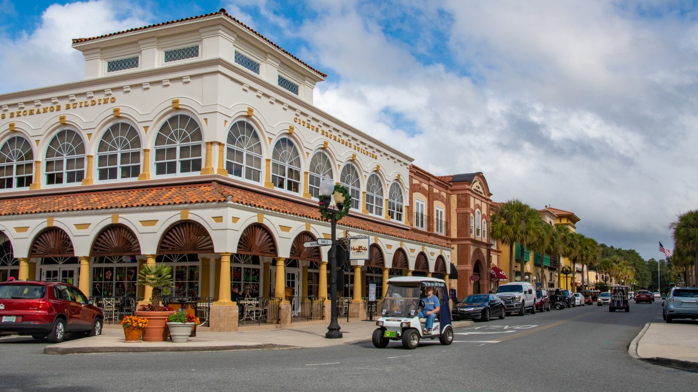 A wide street in The Villages, Florida, with a large, cream-colored building featuring a terracotta roof and arched windows on the left, labeled 'Citrus Exchange Building.' Several golf carts and cars are visible on the road and parked along the curb. Palm trees line the street, and other colorful buildings can be seen in the distance under a partly cloudy sky. A person is driving a white golf cart in the foreground.