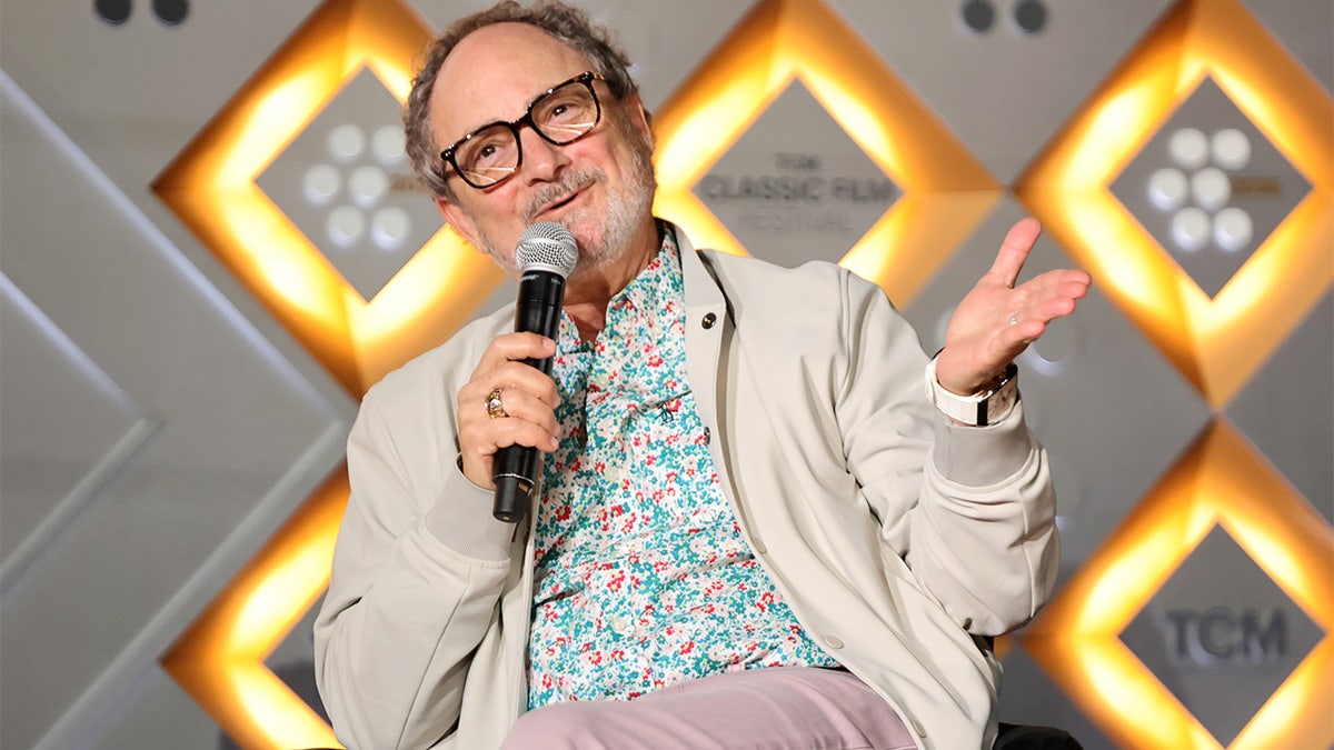 Kevin Pollak wearing glasses and a light jacket speaks into a microphone while gesturing with one hand.
