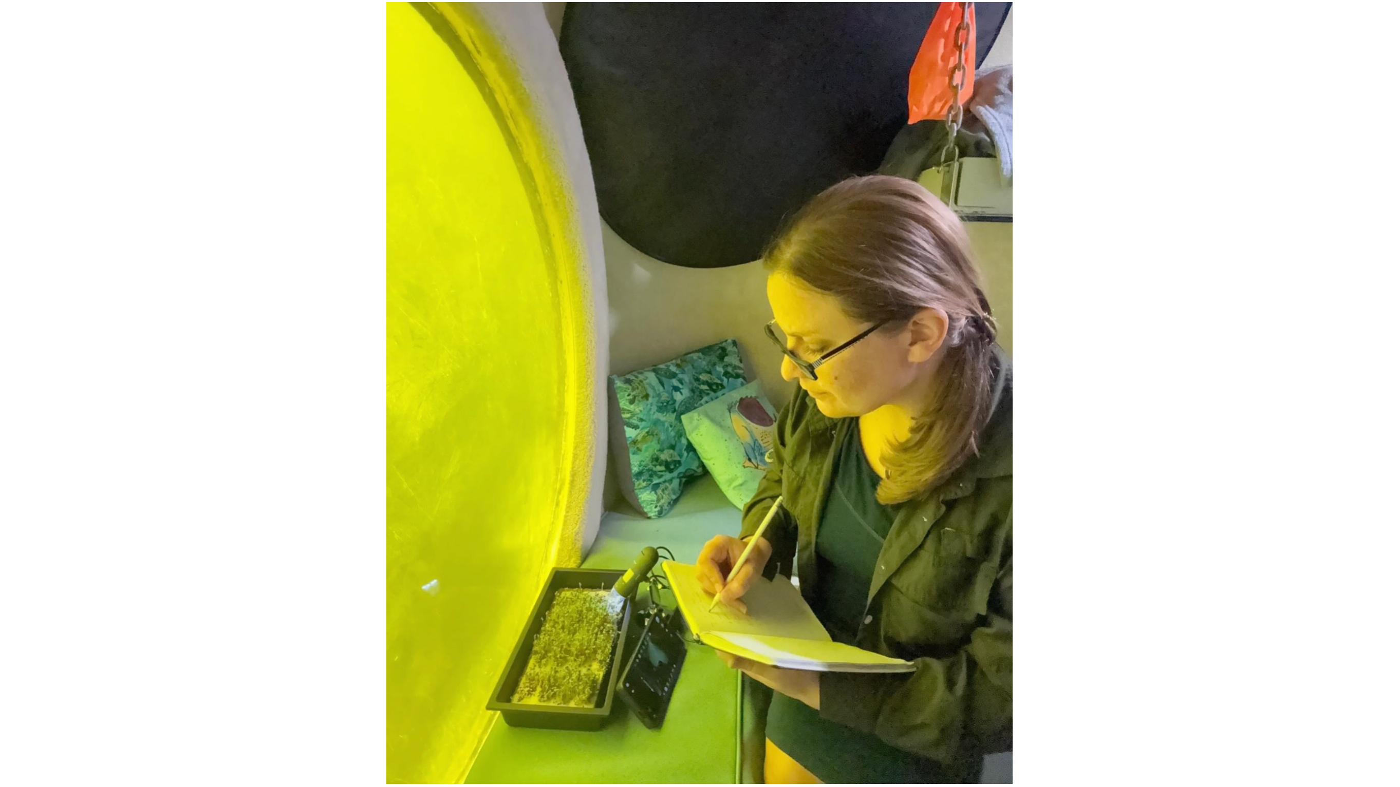 a woman makes notes about the growth of plants inside an underwater research station, next to a circular window looking out into greenish water