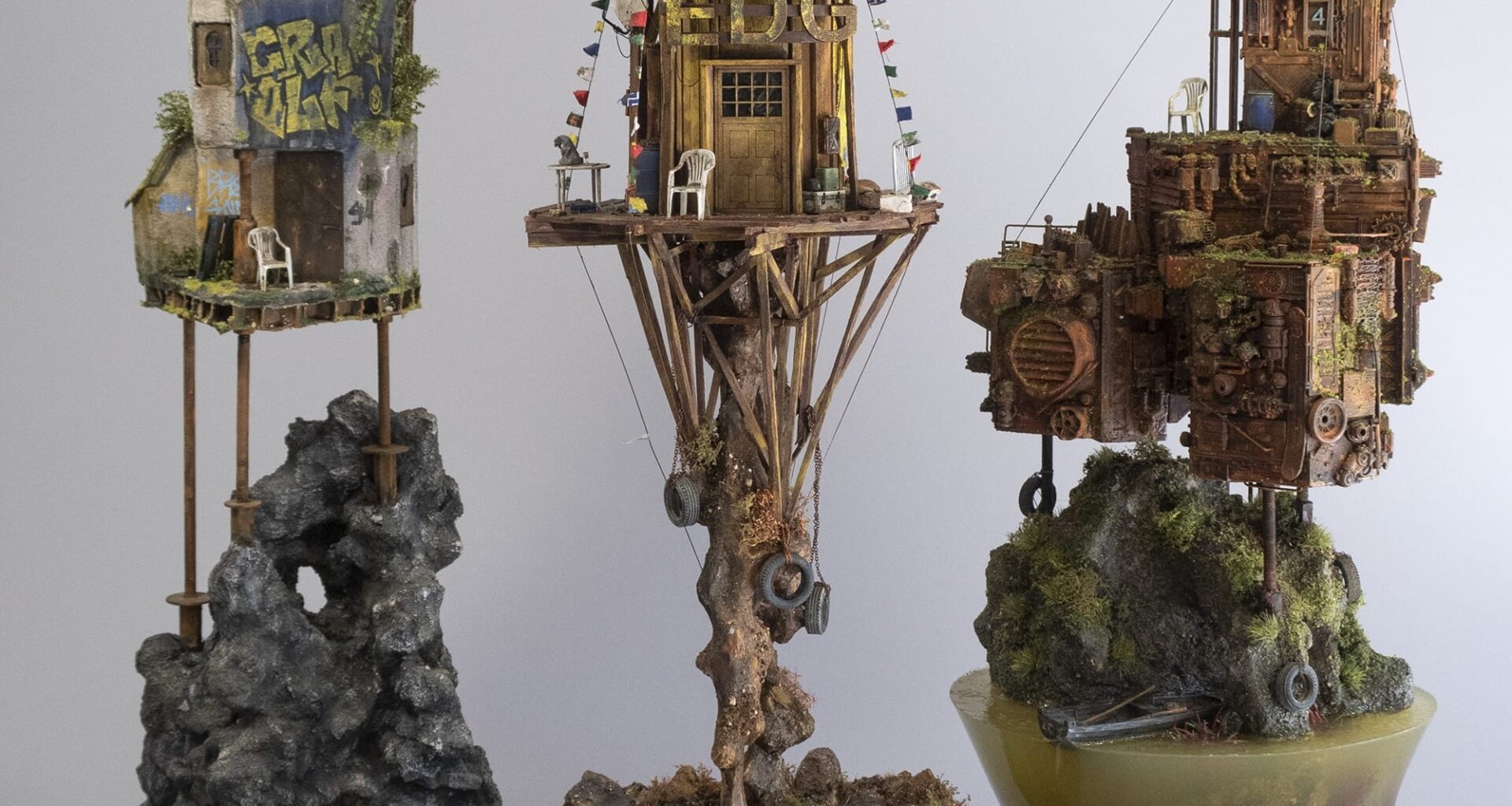 three sculptures by Simon Laveuve in 1/35 scale of ramshackle, dystopian houses built on stilts and rocky outcrops