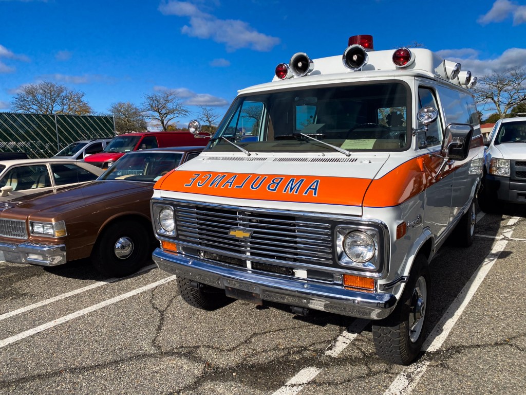 A white and orange vintage Chevrolet van-based ambulance parked on a film set, surrounded by other period-accurate cars.