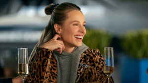 Kristen Bell sits at a table, champagne glasses in front of her, smiling while she looks to her right