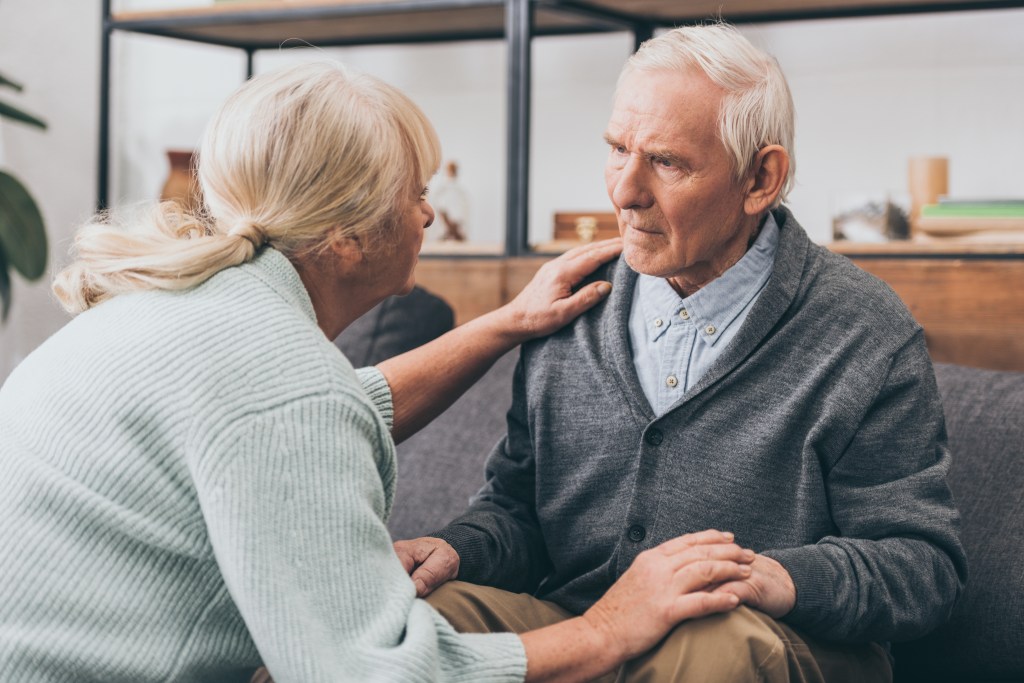An elderly woman holding hands and comforting her upset husband at home.
