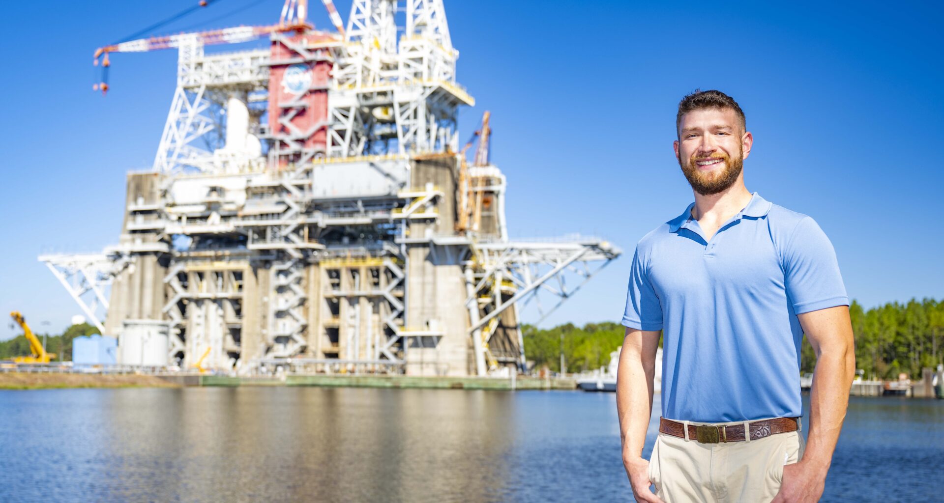 Jared Spiers, an electrical engineering technician at NASA’s Stennis Space Center, stands outdoors and poses for a portrait with the Thad Cochran Test Stand visible in the background.