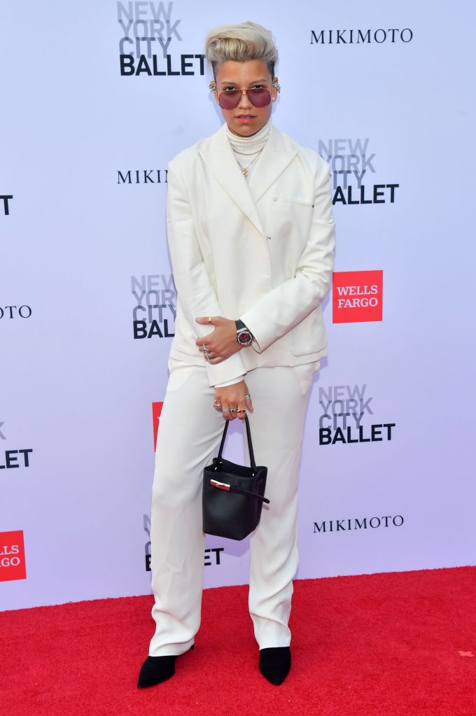 Victoria Brito poses on the red carpet wearing an all-white suit, tinted sunglasses, and holding a small black handbag.