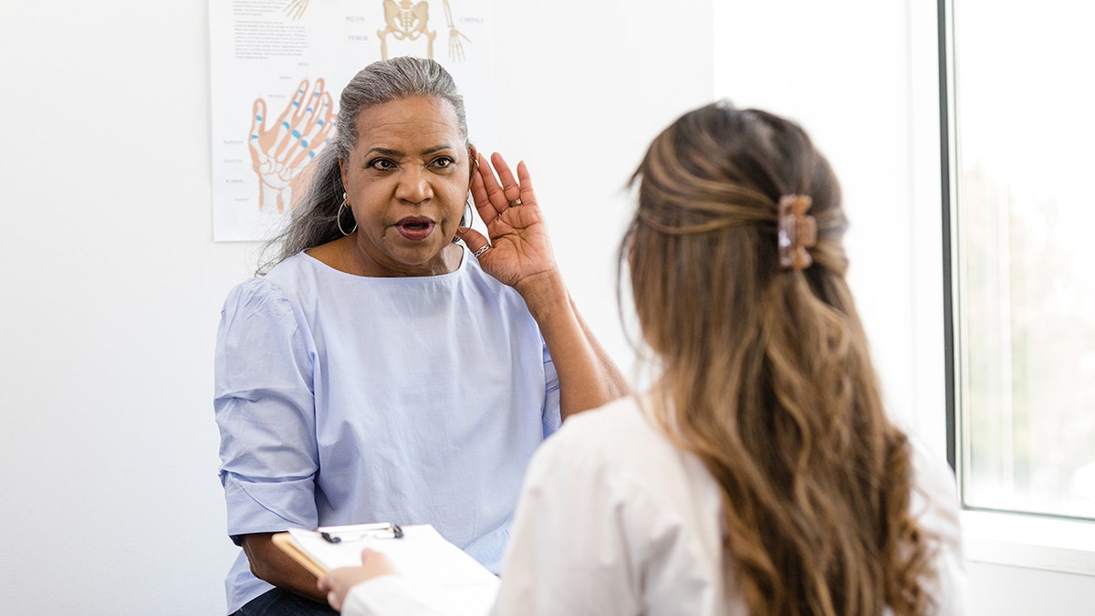While the doctor is talking, a woman places her hand to her ear to let her know that she can't hear very well.