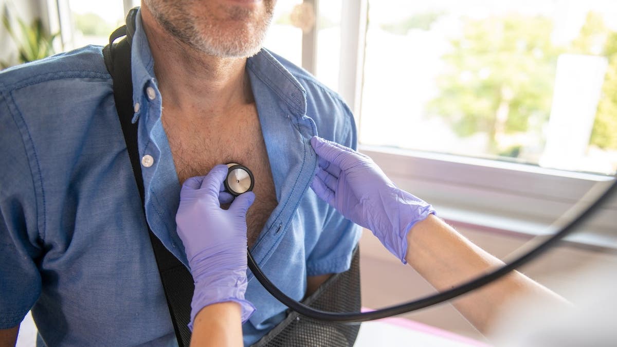 Woman doctor listens to male patient's heart and lungs with stethoscope during medical examination.