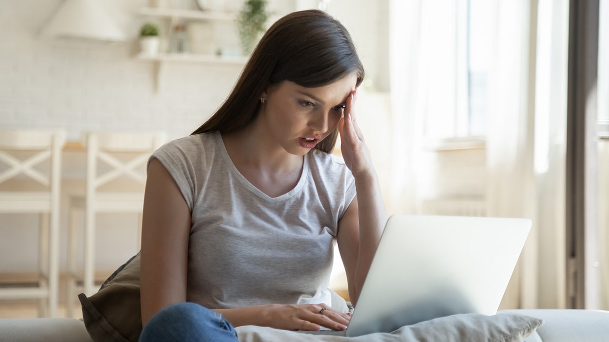 Anxious young woman sitting on couch looking at laptop screen