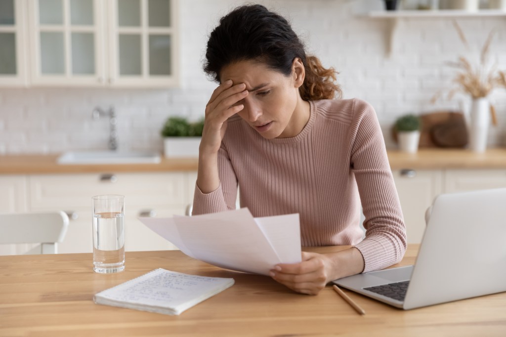A stressed woman sits at a wooden table in her kitchen, holding a piece of paper and looking distressed while reviewing financial documents near a laptop.