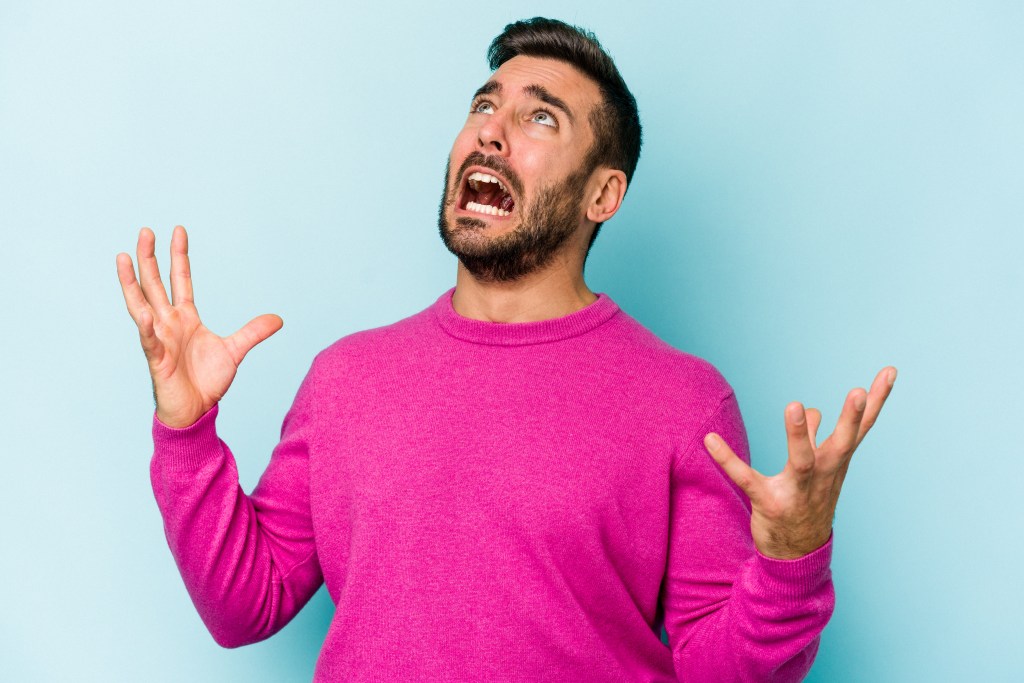 A young man in a pink sweater looks up and screams in frustration, with his hands raised in exasperation against a blue background.