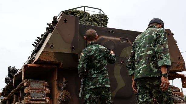 Two men in camouflage military uniforms stand next to a brown military vehicle