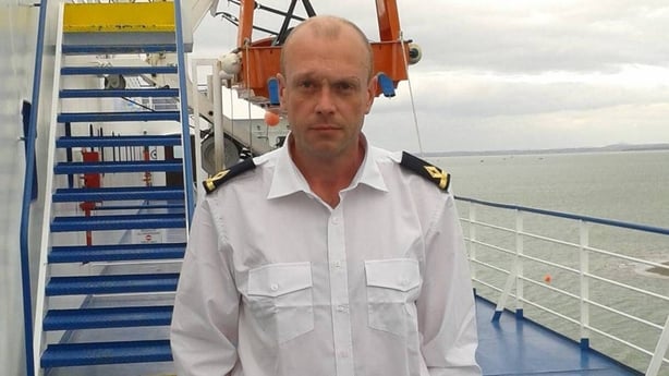 A bald man in a white looks at the camera whilst on a boat with a blue deck with the sea in the background