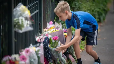 Getty Images A boy wearing a blue shirt and black shorts is laying flowers on the ground next to other bouquets. They are by a school fence.