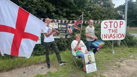 Richard Knights/BBC Picture shows three men, one holding a St George Flag and the other is sitting down while holding a poster. The third man is standing next to a sign that reads 'Stop the invasion'.