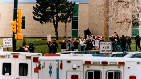 Getty Images More than a dozen students are given an armed escort from Columbine High School in Colorado, US, after two teenagers went on a shooting spree. The pupils, photographed from a distance are walking in a straight line with their hands on their heads. Two white "Right Lane Must Turn Right" signs are visible and a line  ambulances are parked alum the pavement.  