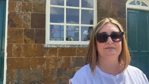 Annabel Amos/BBC A woman wearing sunglasses in front of a stone building with a green door.