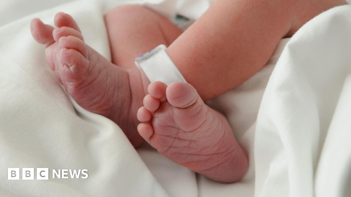 Picture of a newborn baby's naked feet, which are crossed at the ankle. There is a plastic identification tag on one ankle and is laying on a white cotton sheet.