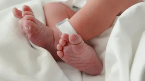 Getty Images Picture of a newborn baby's naked feet, which are crossed at the ankle. There is a plastic identification tag on one ankle and is laying on a white cotton sheet.