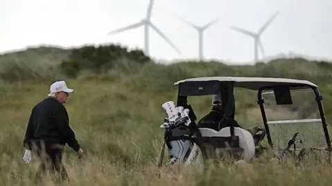 Getty Images President Trump in a white cap walks towards a golf buggy with wind turbines in the distance