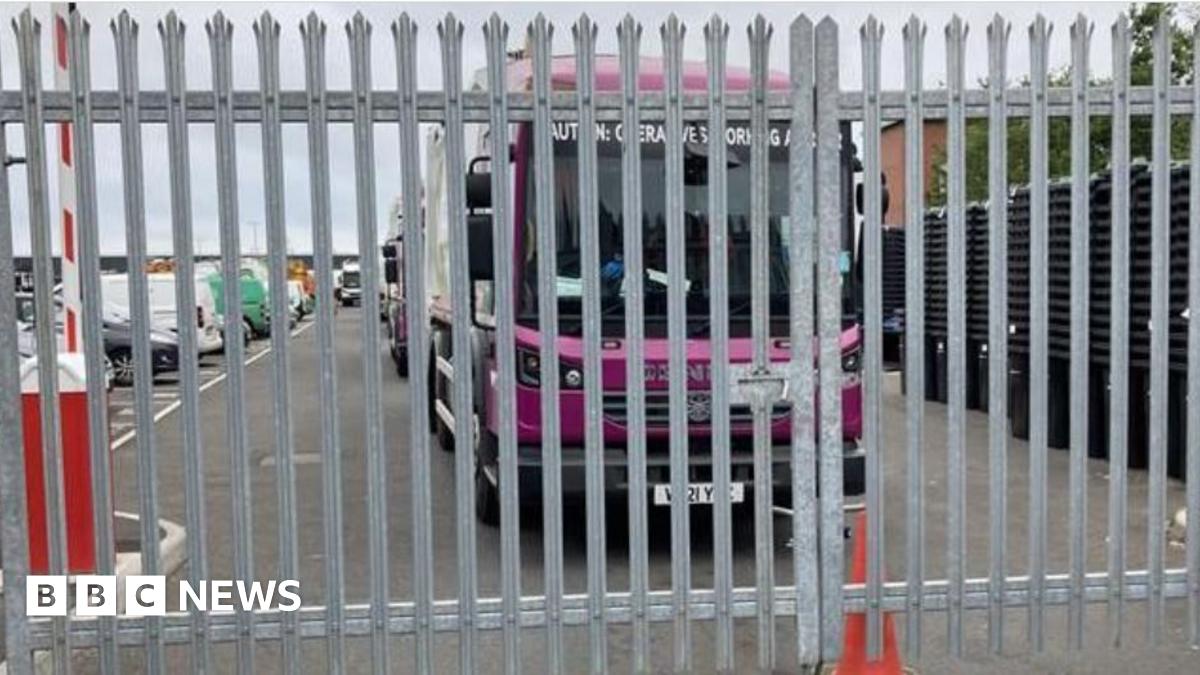 A purple waste lorry sits behind metal gates. An orange cone can be seen in front.