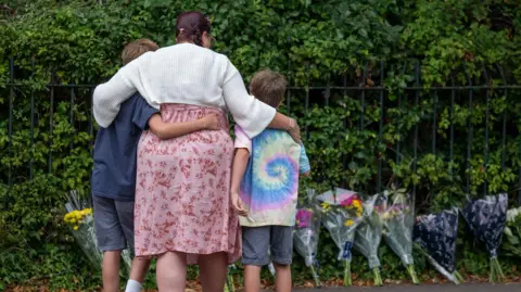 Getty Images A woman, who wears a white cardigan and a pink floral dress, holds her arms around two boys. One on the left is taller, and wears a navy t-shirt and grey shorts. The boy on the right wears a tie-dye t-shirt and grey shirt. They are photographed from behind, as they look at a row of floral tributes against a hedge and a fence. 