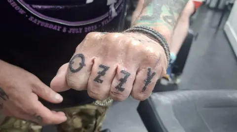Simon Humphreys A man posing with his arm toward the camera. He is wearing a black and purple Black Sabbath concert t shirt. His hand is close to the camera, with a closed fist showing his Ozzy tattoo across his four fingers