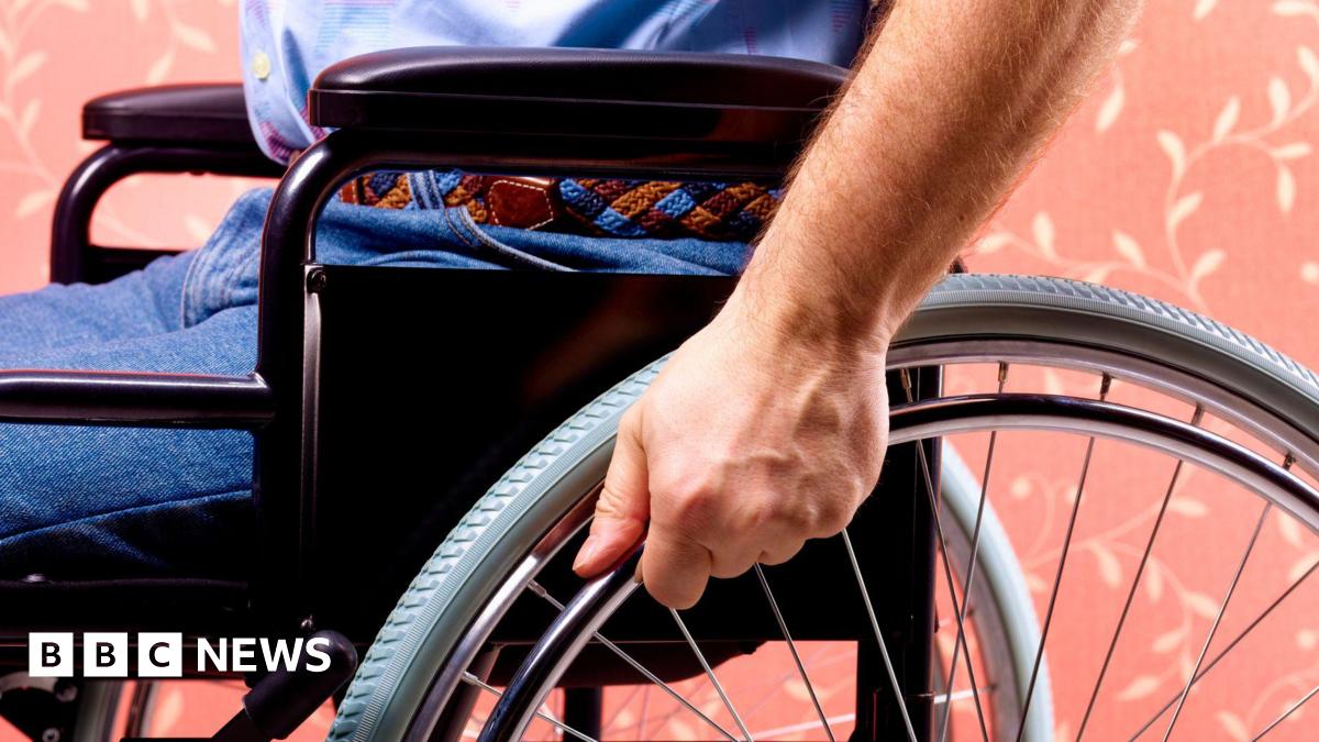 Close-up of an anonymous man sitting in a wheelchair. He is gripping one of the wheels with his left hand. He is wearing belted blue jeans and a light blue shirt.