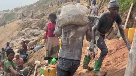 Hassan Lali / BBC Muddy workers are seen on a slope at Rubaya mine in DR Congo. One in green wellingtons, black jeans and T-shirt looks at the camera, another in a similar outfit is seen from the back as he walks with a sack on his shoulders. A group of women are seen a little further down the slope near some big basins and jerry cans.