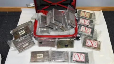 National Crime Agency Piles of drugs in clear wrapping piled in and around a black and red suitcase.
