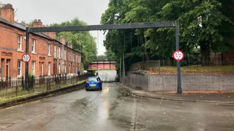 Richard Stead/BBC A blue car drives under a canal bridge on a residential road. People walk along the left side of the road with umbrellas. 