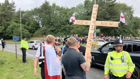Alex Dunlop/BBC A man holding a large wooden cross that reads 'stop the boats now' with two St George flags attached on either sides of them. There are police officers also at the scene 