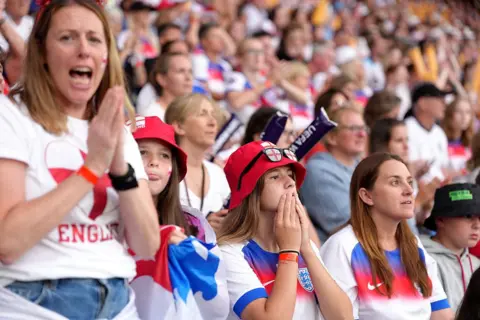 Getty Images Four nervous looking female football fans in England kits watch with hands to their mouths in a packed stadium.