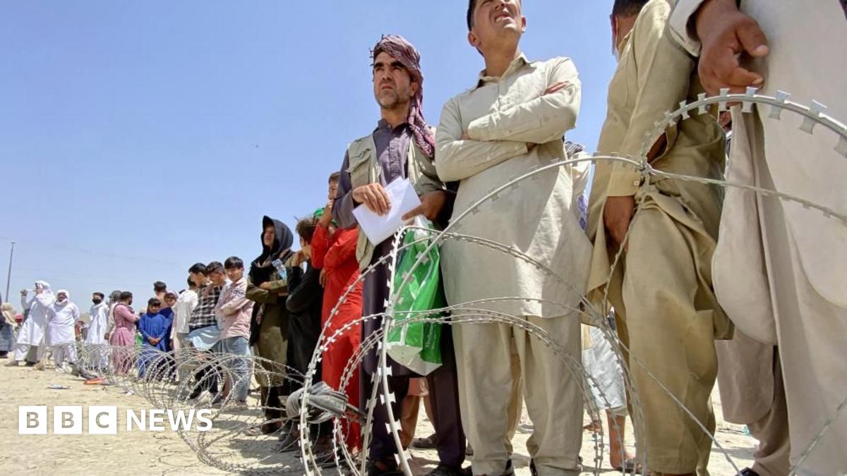 Afghans wait patiently in line in Kabul in 2021 to flee on evacuation flights, some with their arms crossed, with barbed wire in the foreground.
