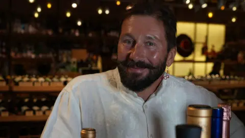 A man with a beard and moustache smiles at the camera. He is in a wine shop with bottles behind him on the shelf