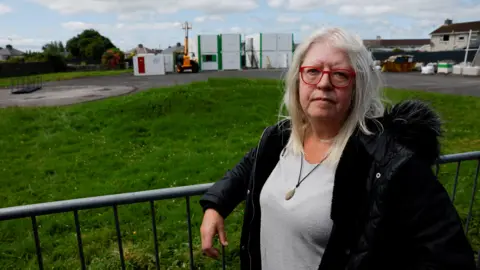 Reuters A woman with white hair and red glasses looks at the camera. She is wearing a grey jumper and a black jacket. She has her arm on a metal fence, and behind her is a patch of grass and a set of buildings. 
