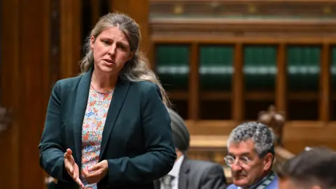 House of Commons Rachael Maskell stands and gestures with her hand as she addresses the House of Commons. She has long, wavy blonde hair and wears a dark green jacket and a floral top. 