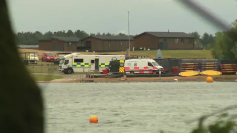 Image shows a lake in the foreground, with a police mobile incident unit on the shore, a search and rescue team van next to it, two crates full of canoes also on the shore, and chalet buildings in the background