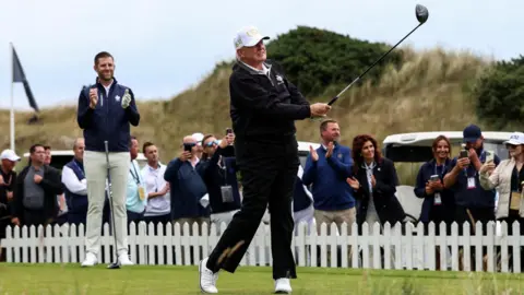 PA Media Donald Trump after taking a golf swing. His son Eric is applauding on the course, along with a number of spectators behind a small, white fence