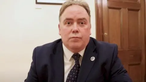 Croydon Council A middle aged man sitting down in an office - he is wearing a dark blue suit, a white shirt and blue patterned tie. A wooden door is visible behind him.